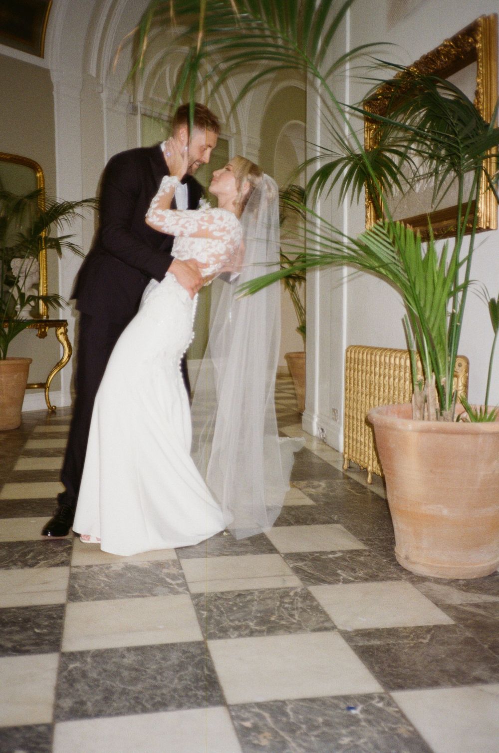 A romantic wedding couple shares a tender dance moment in an elegant hallway adorned with palm plants and marble floors.