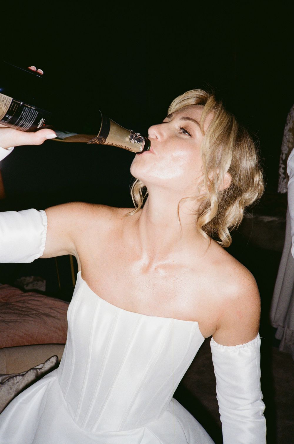 Person in white strapless wedding dress and long gloves drinking from a champagne bottle at a celebration.