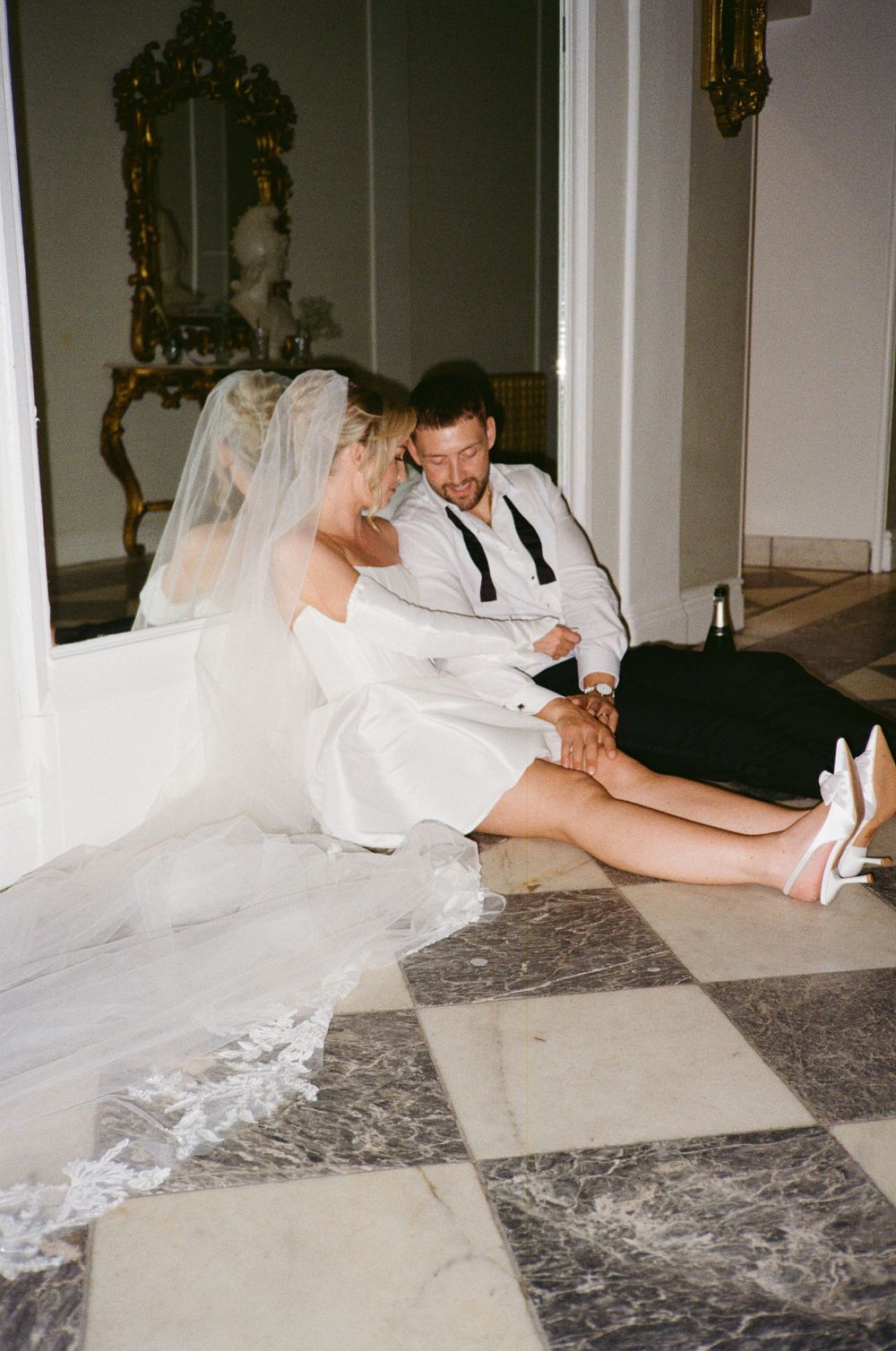 A bride in a white wedding dress and veil lays on the floor in a playful pose at an indoor venue.