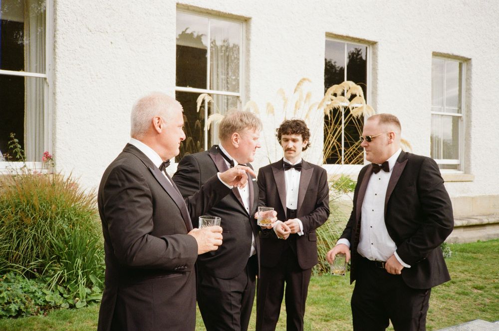 A group of formally dressed men in suits and tuxedos gather outside a white building during a wedding.