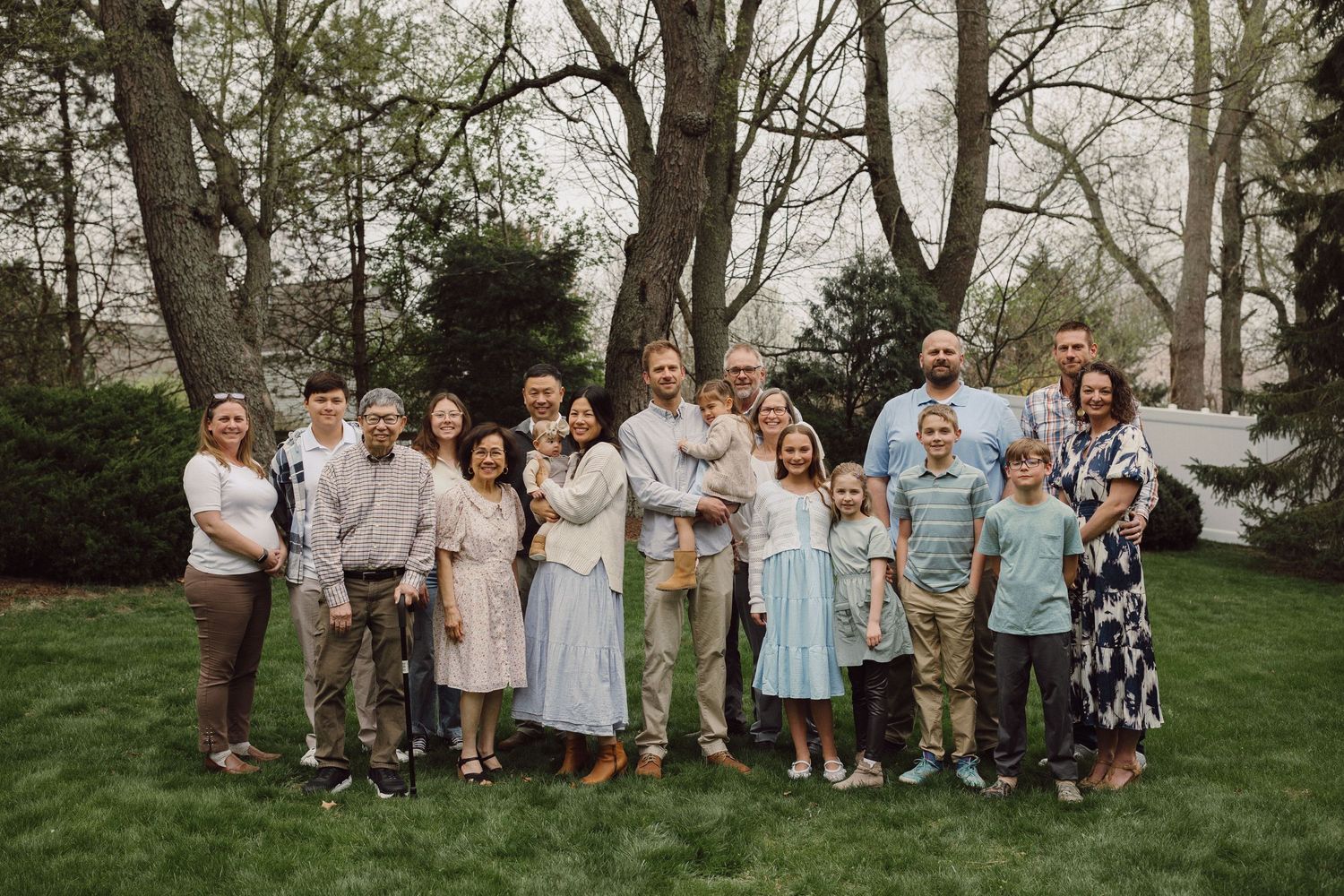 Family group photo outdoors in casual attire standing together on a lawn with trees in background.