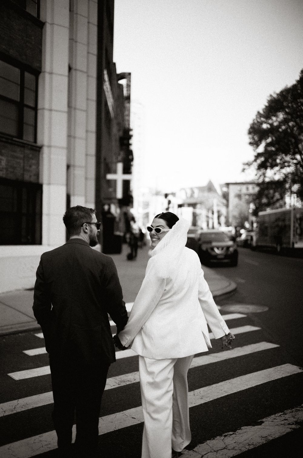 A couple in wedding attire walking across an urban crosswalk in black and white.