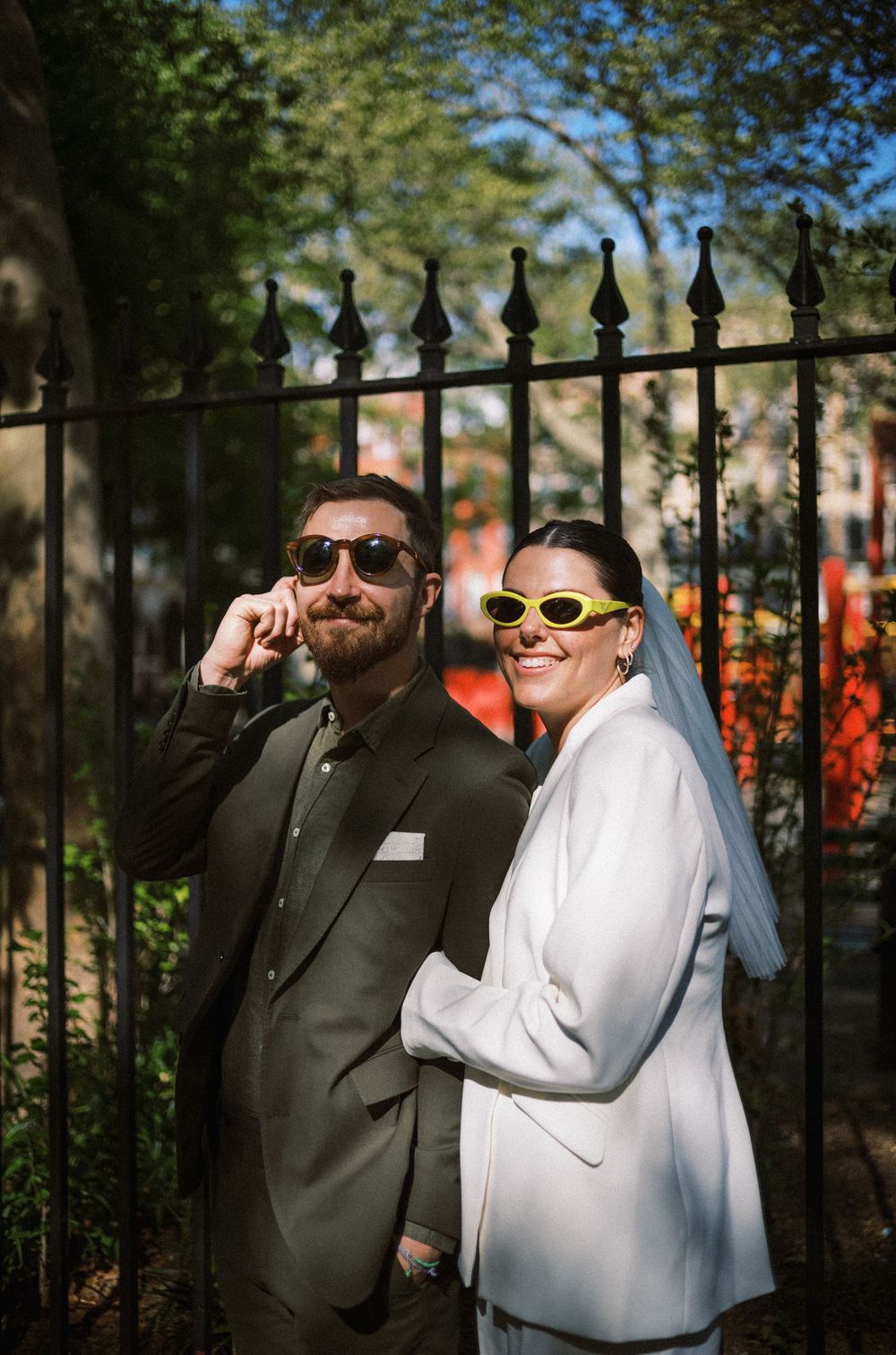A stylish couple in sunglasses posing against an iron fence on their wedding day.