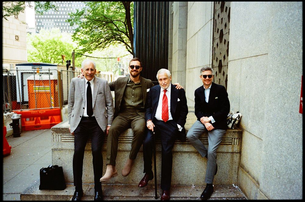 Four people in formal attire sit casually on a stone ledge outside a building on a sunny day.
