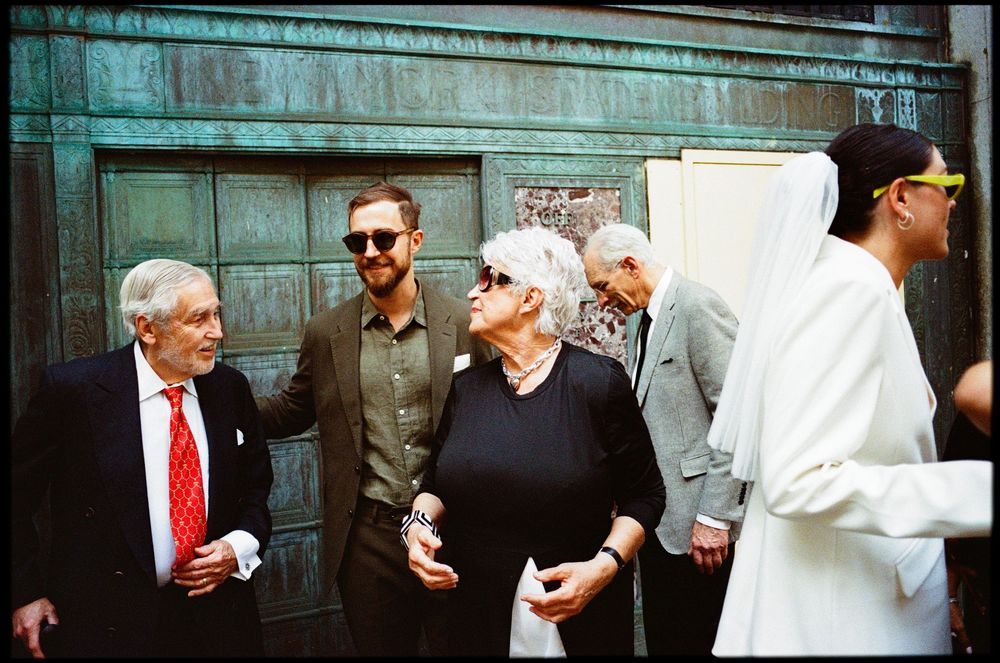 A group of people in formal attire stand together conversing next to a dark wooden door with a greenish tint.