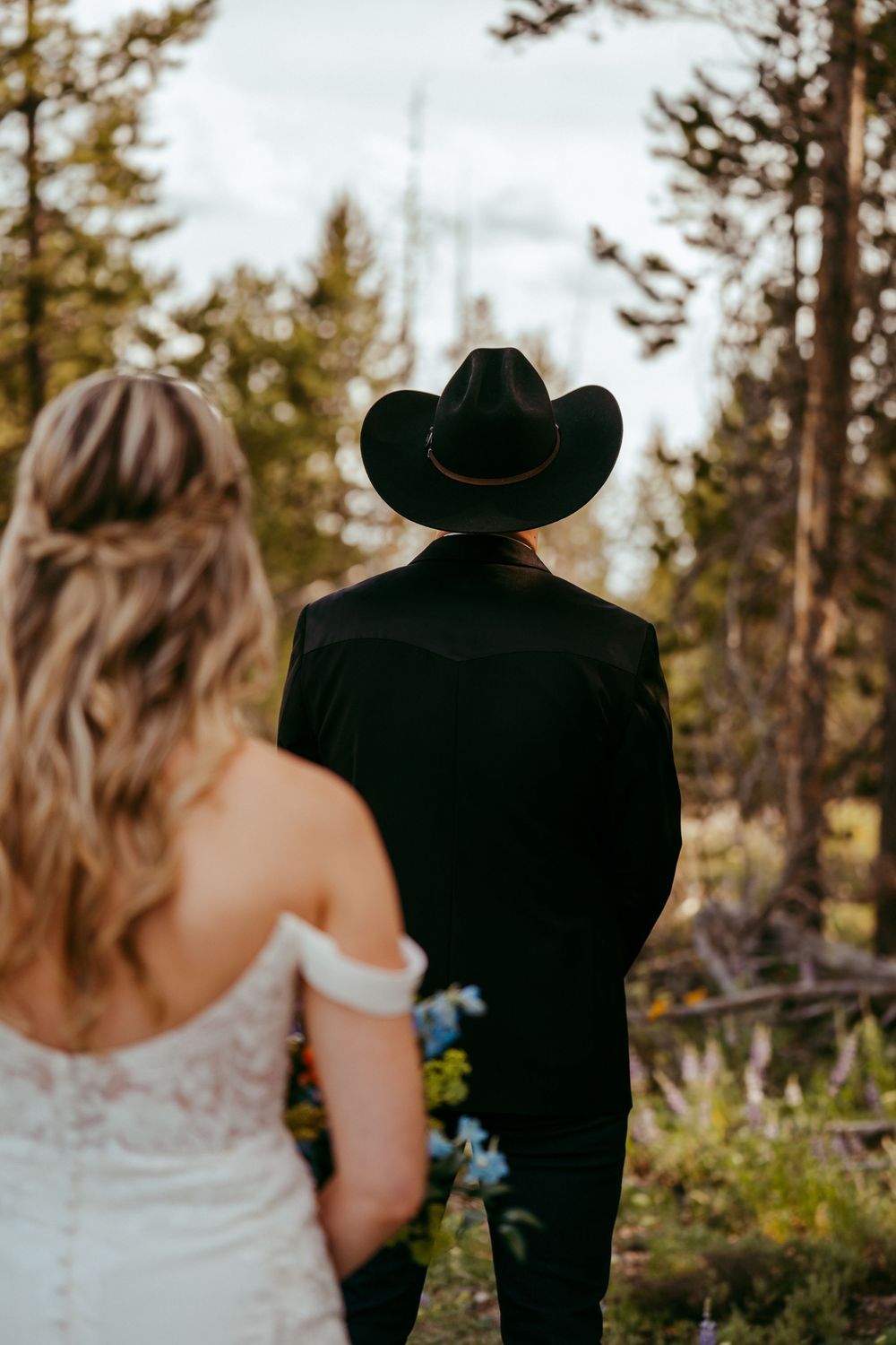 Rustic mountain wedding ceremony with a bride and groom exchanging vows in a wildflower meadow, surrounded by pine trees and mountains