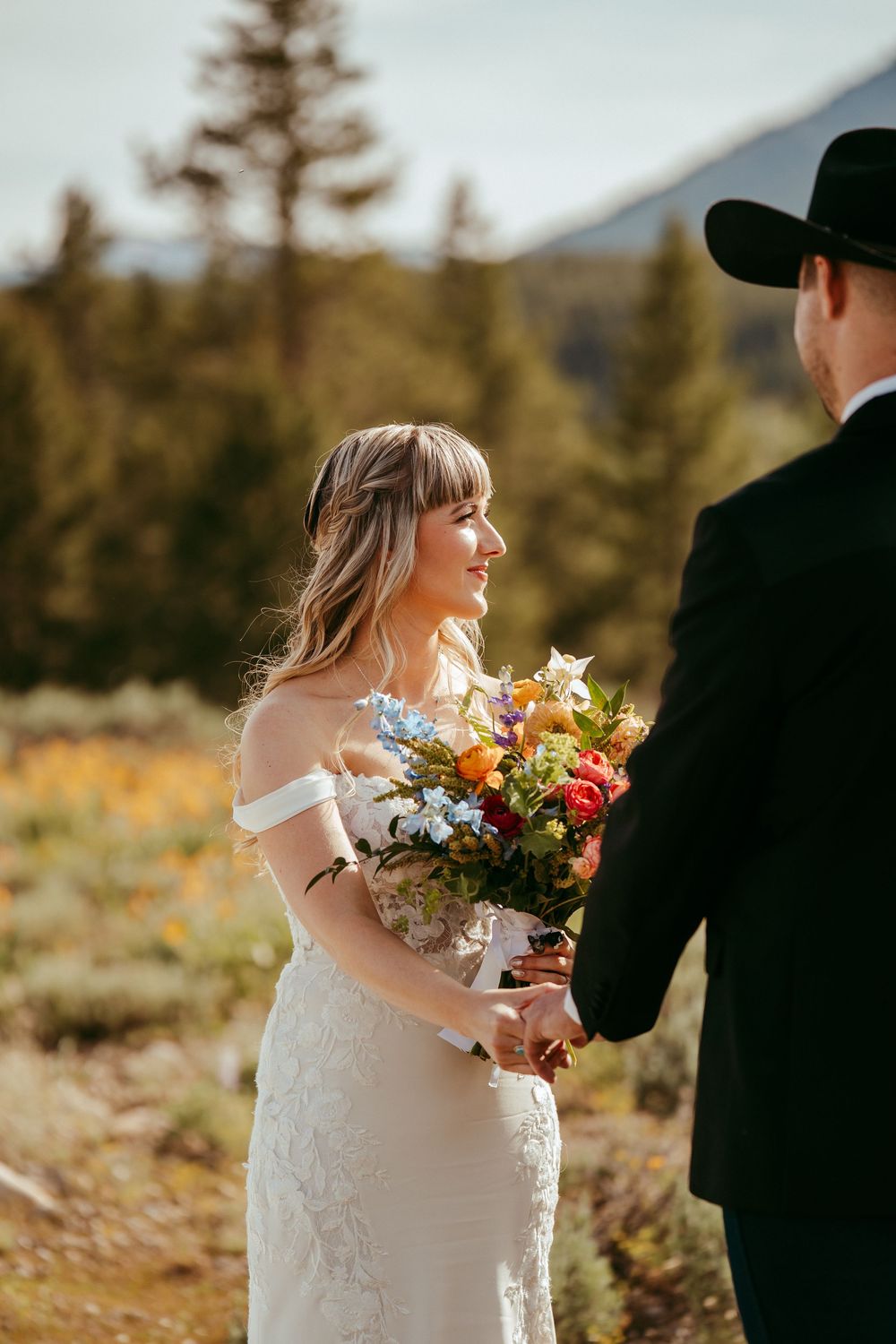 Rustic mountain wedding ceremony with a bride and groom exchanging vows in a wildflower meadow, surrounded by pine trees and mountains
