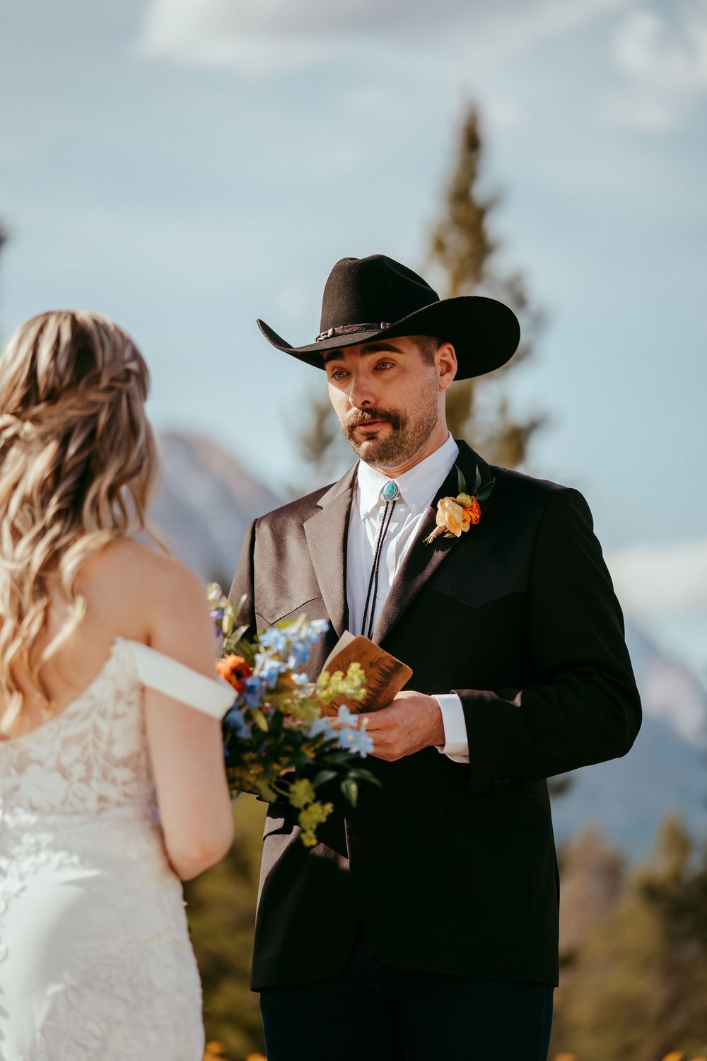 Rustic mountain wedding ceremony with a bride and groom exchanging vows in a wildflower meadow, surrounded by pine trees and mountains