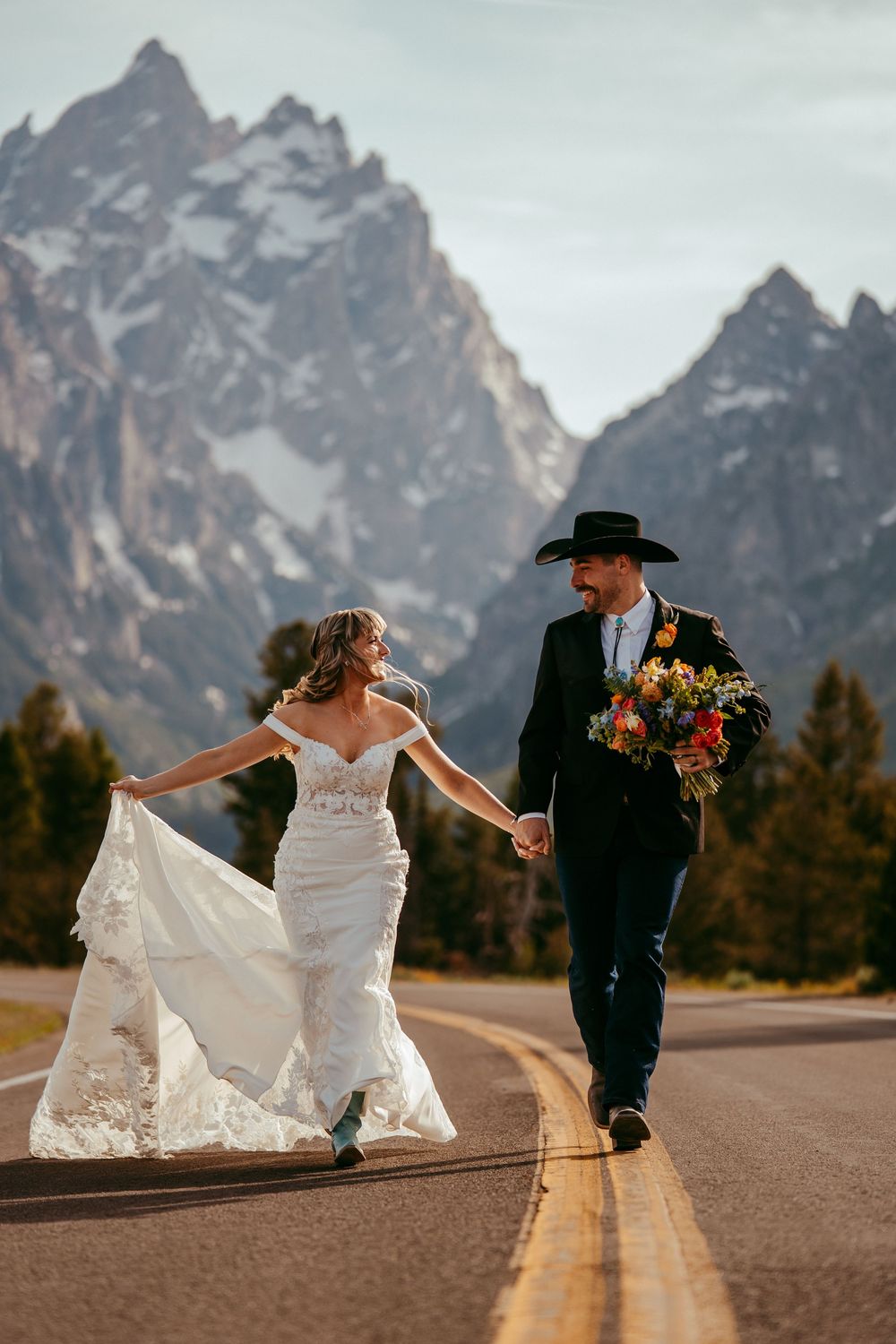 Joyful western wedding photo of a bride and groom running hand-in-hand down a scenic mountain road