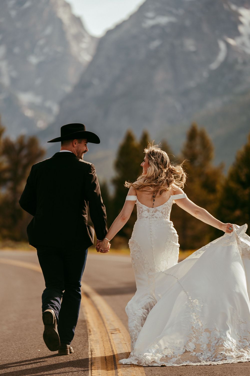 Joyful western wedding photo of a bride and groom running hand-in-hand down a scenic mountain road