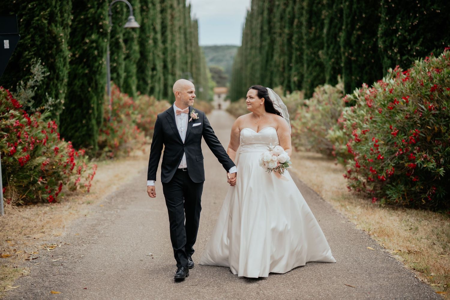 A couple in wedding attire walks hand in hand down a cypress tree-lined path with red flowering bushes in Italy.