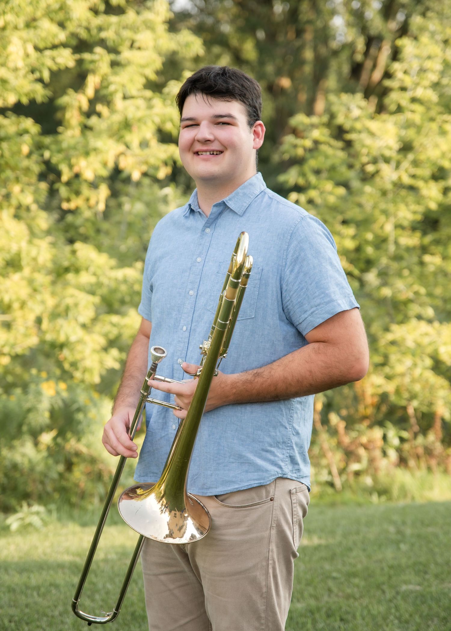 Student musician poses in khaki pants and light blue shirt while holding trombone in sunny outdoor setting.