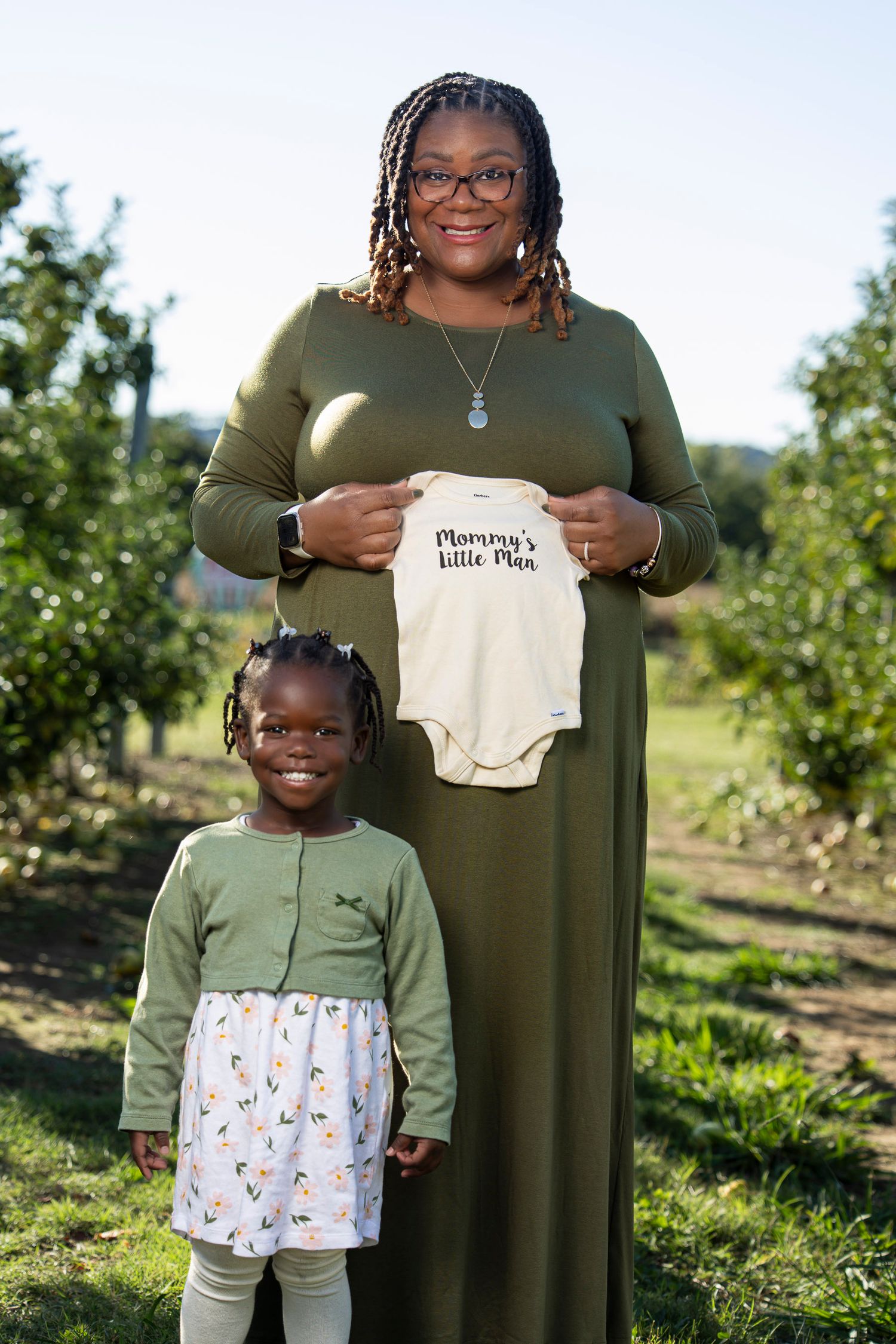 A parent and child in matching olive green outfits pose together in Rivercrest orchard on a sunny day in Fayetteville AR