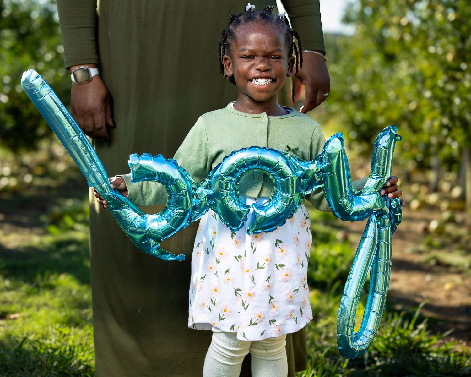 A person holds teal balloon letters spelling "boy" while standing in a lush green garden setting at Rivercrest Orchard in Fayetteville AR.
