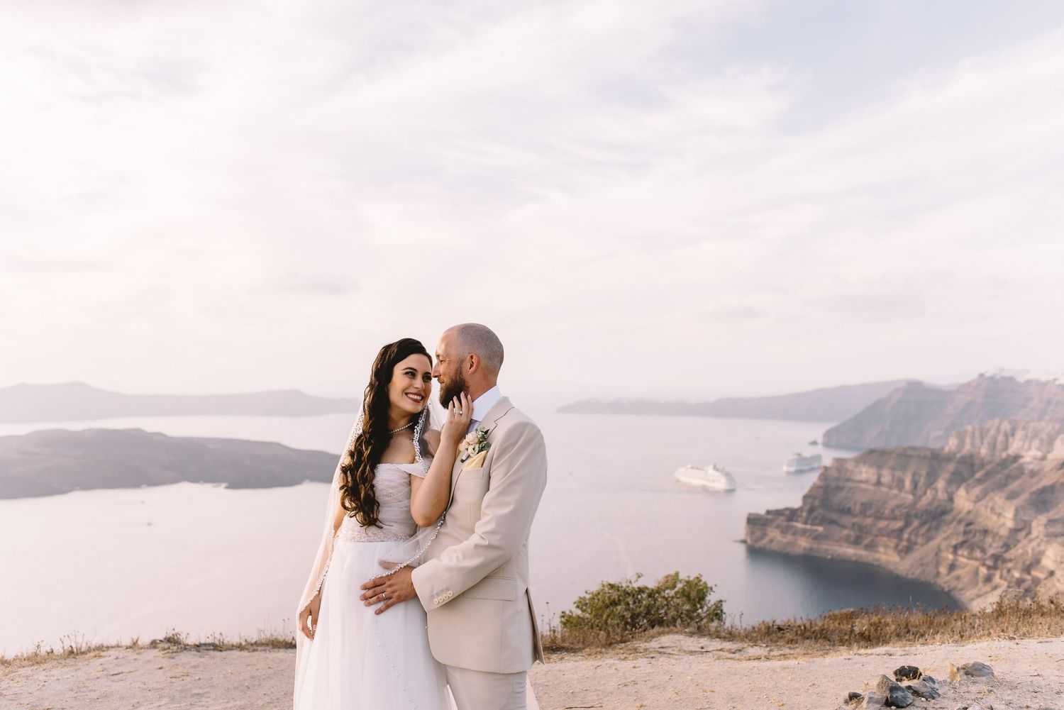 Wedding couple embraces on scenic clifftop overlooking Mediterranean coastal waters during sunset.