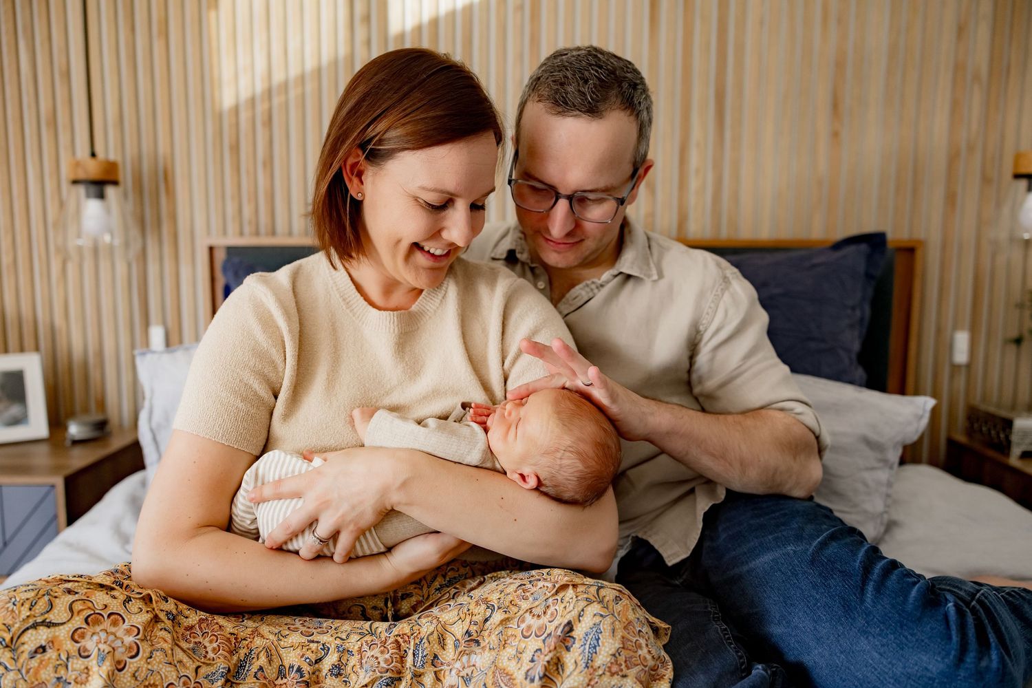 Parents sharing an intimate moment with their newborn baby on a bed with a floral patterned blanket.
