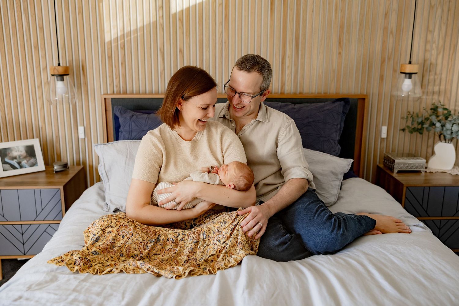 A couple rests and snuggles together intimately on a bed with a textured golden knit blanket in a wood-paneled room.
