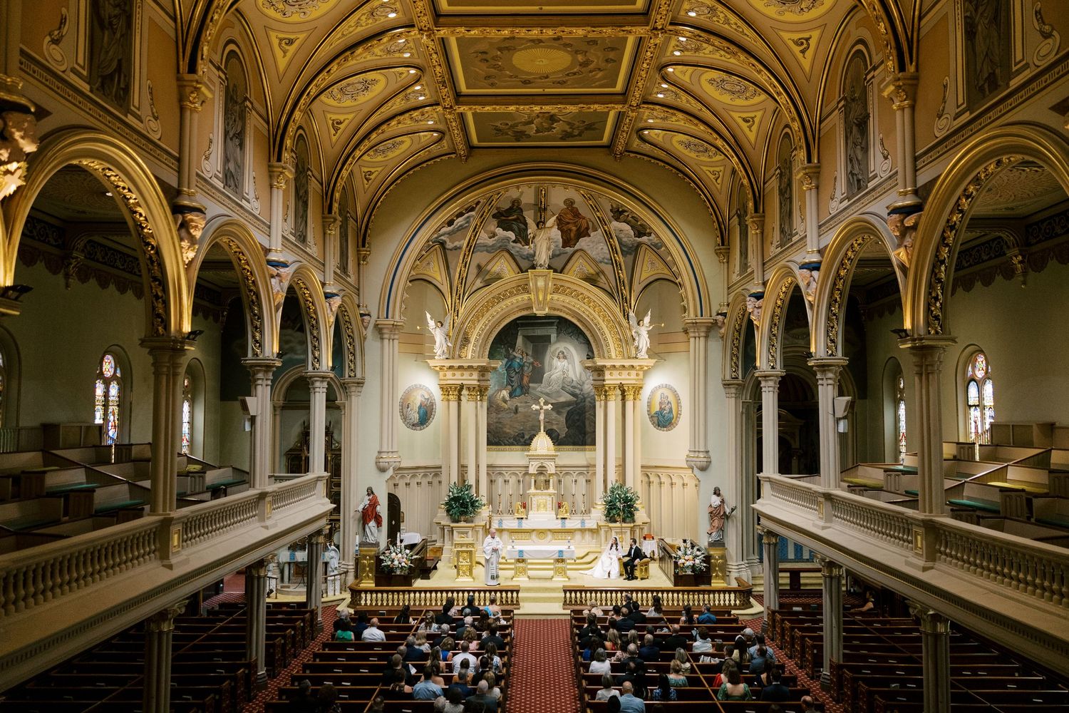 A wedding ceremony taking place inside a stone chapel with vaulted wooden ceilings and a red carpet aisle.