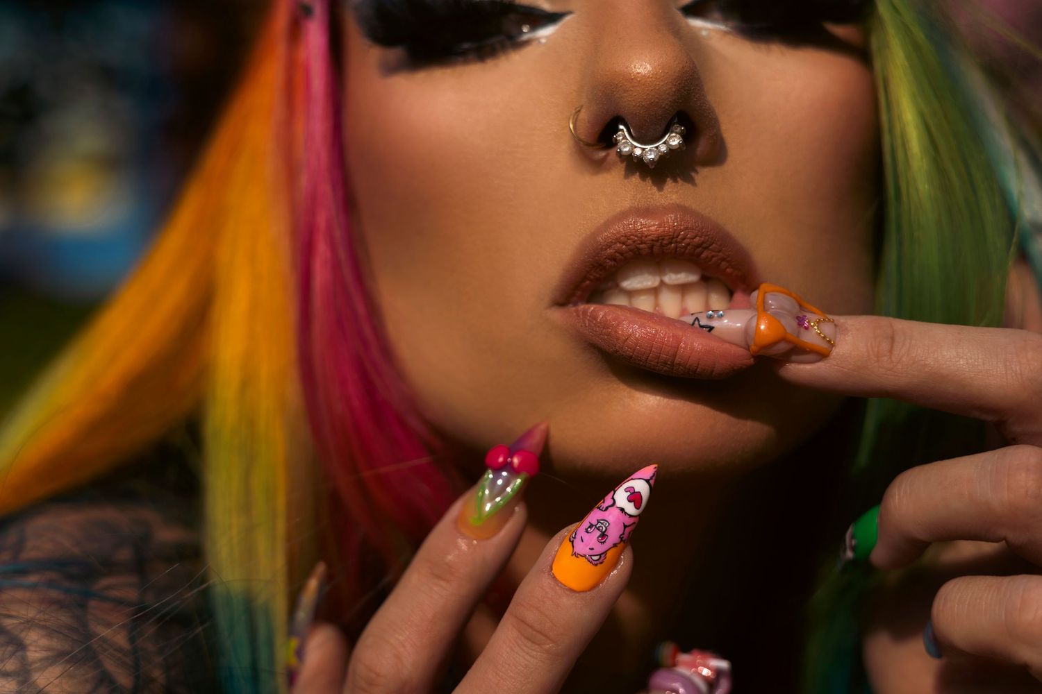 Colorful manicured nails and a septum piercing shown in closeup with rainbow-colored hair visible.