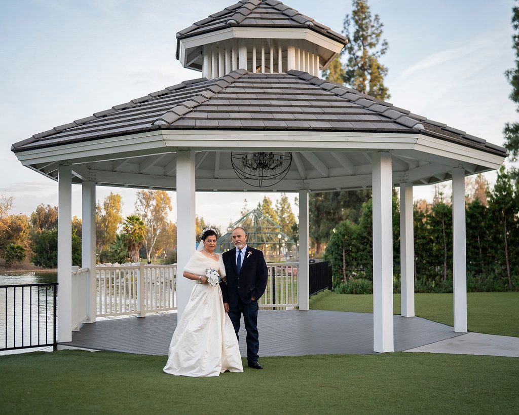 Bride and groom pose together under a white gazebo beside a lake and lawn.