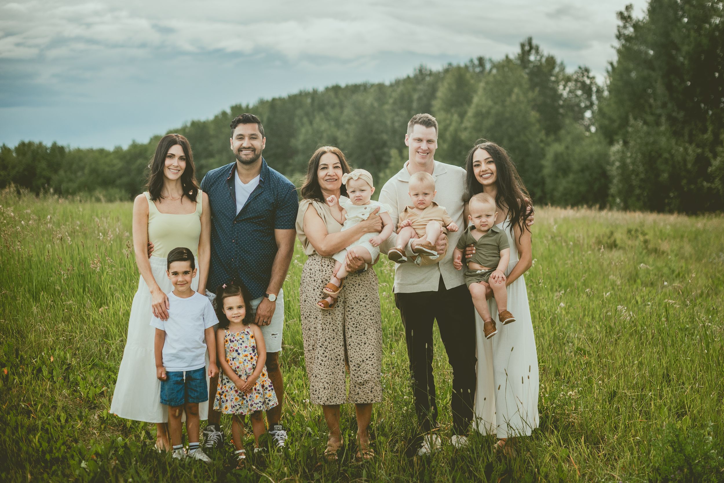 A group poses together in a grassy meadow with rolling hills and trees in the background on an overcast day. St Albert Family Photographer