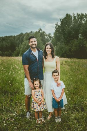 A family poses together in a grassy meadow with trees in the background on an overcast summer day. St Albert Family Photographer