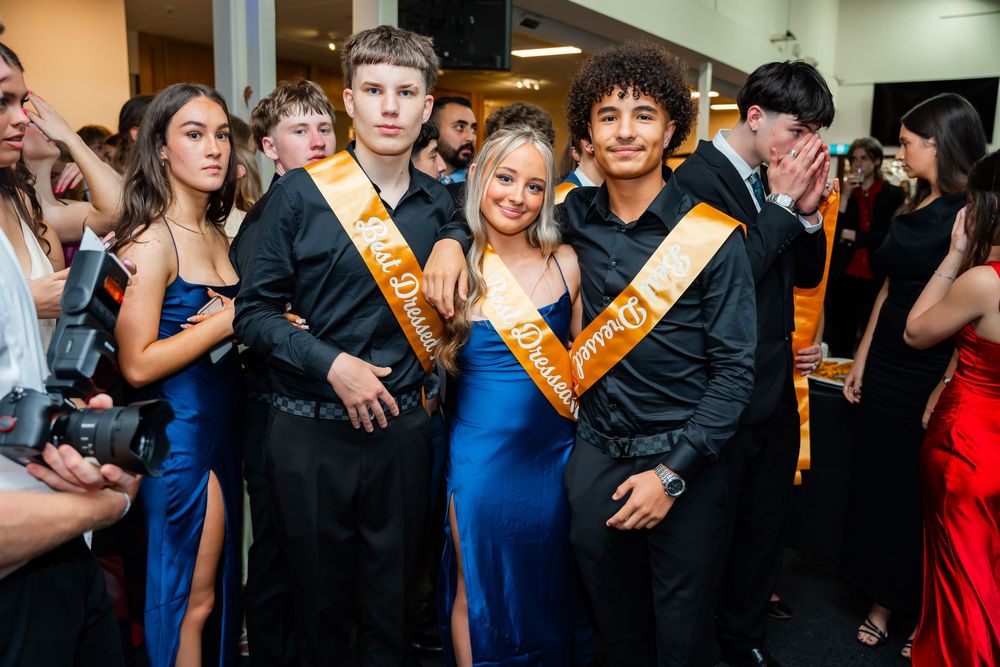 Dancing couples in formal attire move across a dance floor at a celebratory event with orange sashes visible.