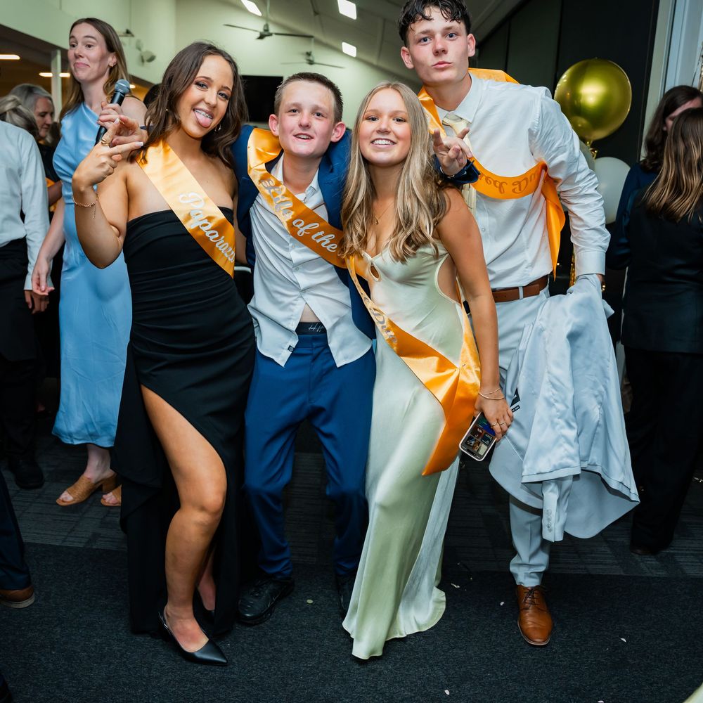 Celebratory group wearing orange sashes and formal attire pose together at an indoor party venue.