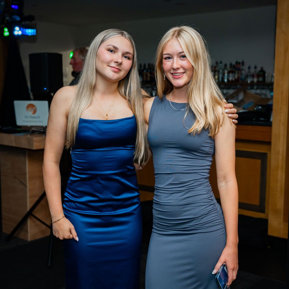 Two women in blue and gray formal dresses smile and pose together at a social gathering.