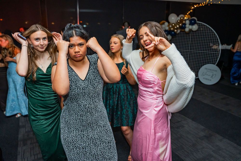 A group of women in formal dresses dance and pose together at a party on a dark dance floor.