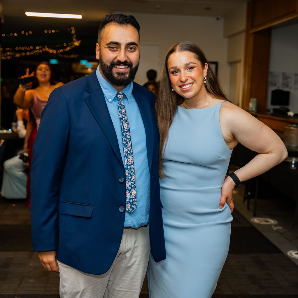 A pair of attendees at an evening event pose together, with one in a blue blazer and tie and another in a blue dress.