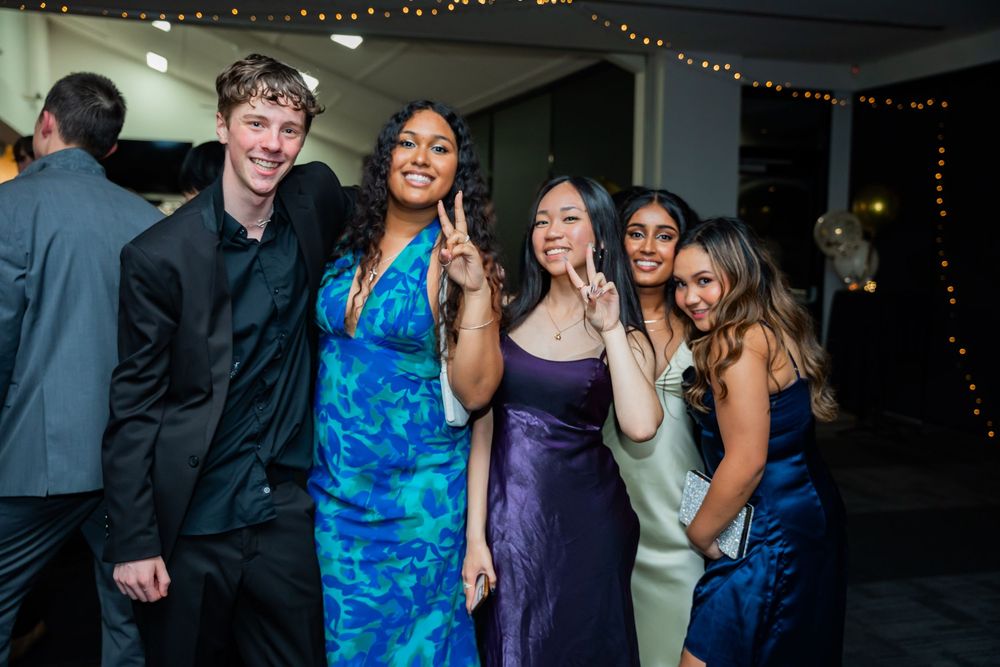 A group of young people in formal attire pose together at an evening event with festive string lights in the background.