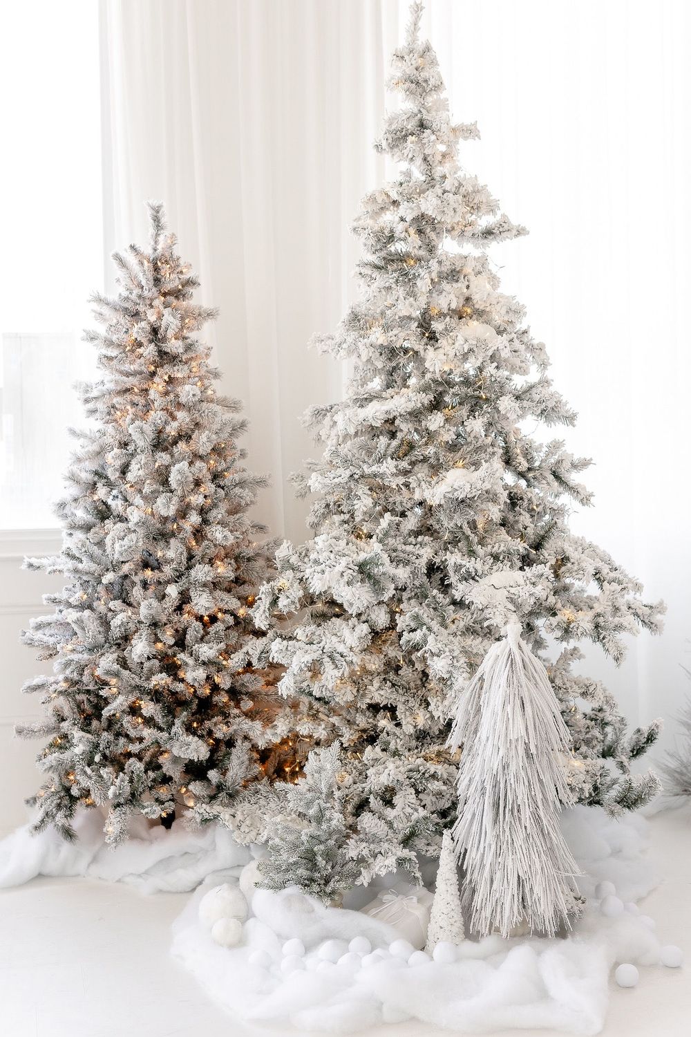 Two flocked artificial Christmas trees decorated with lights and yarn tassel ornaments in a bright white room.