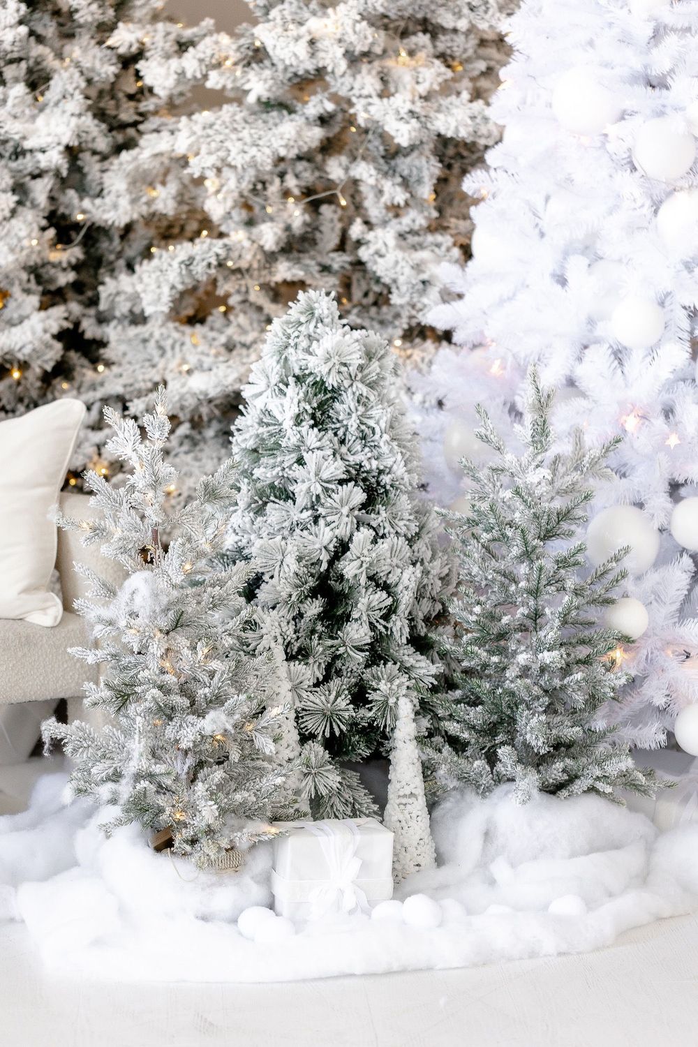Frosted artificial Christmas trees with white snow decor stand against a backdrop of twinkling lights.
