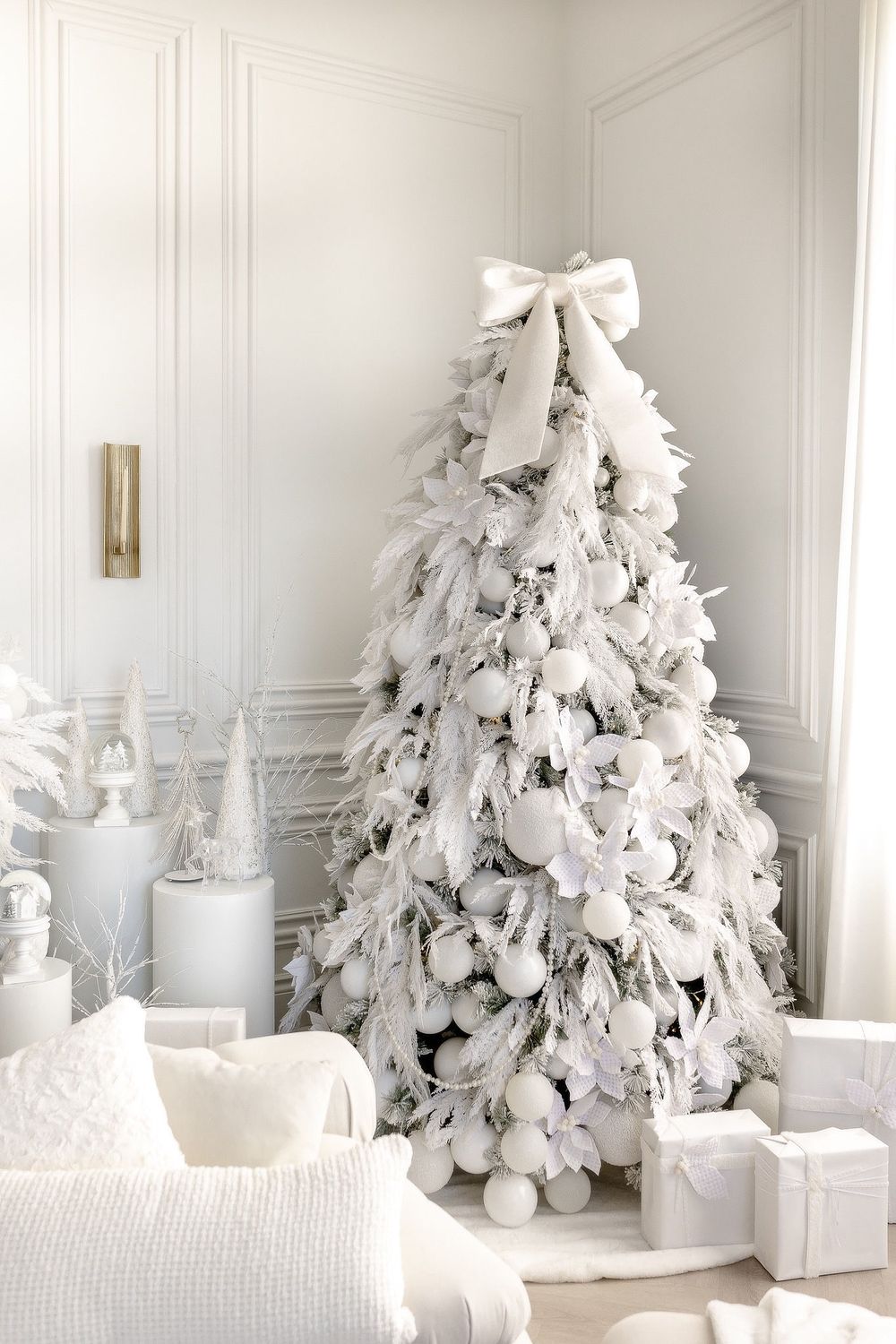 Elegant white flocked Christmas tree decorated with a large bow topper and coordinating presents in an all-white room setting.