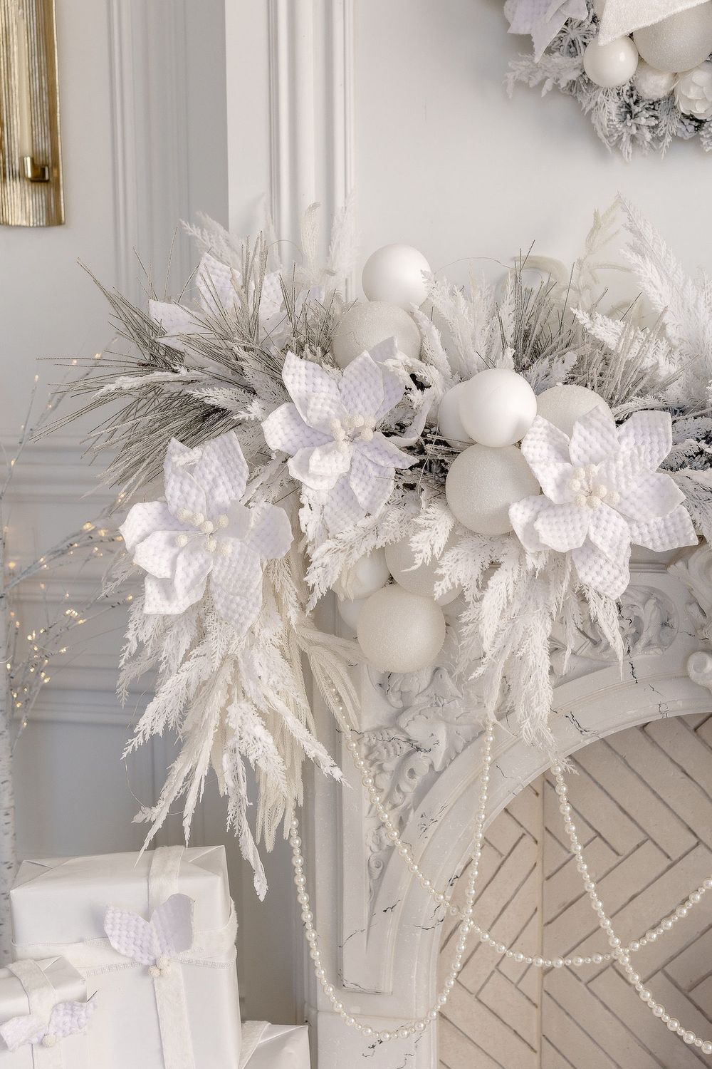 Elegant white Christmas wreath with faux poinsettias, frosted branches, pearl beads and white baubles on a light wall.