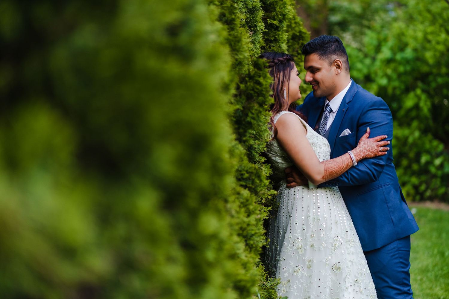 Romantic wedding photo of couple embracing by green hedge in formal attire.