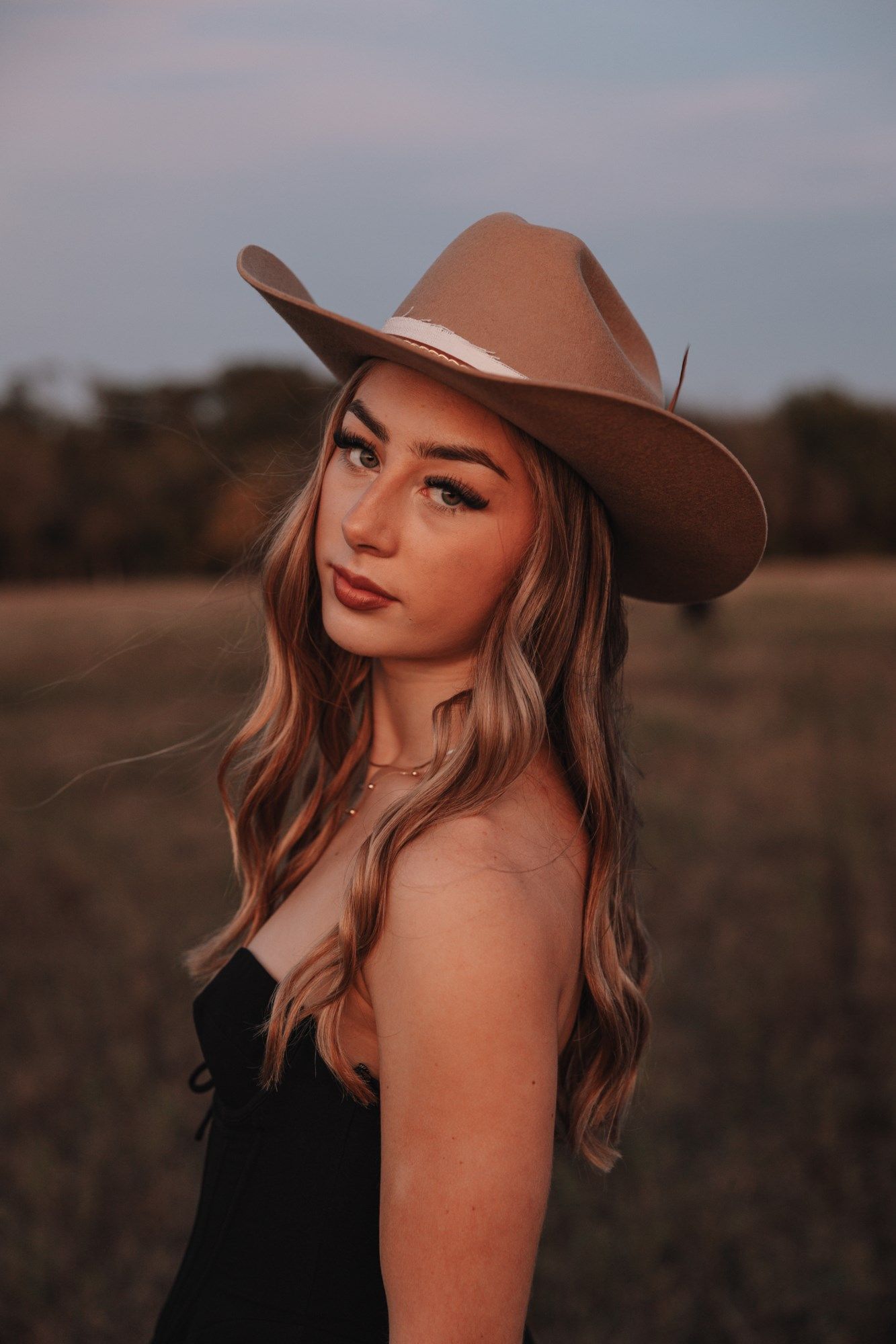 Woman in black top and brown hat poses against sunset backdrop in rustic outdoor setting at golden hour.