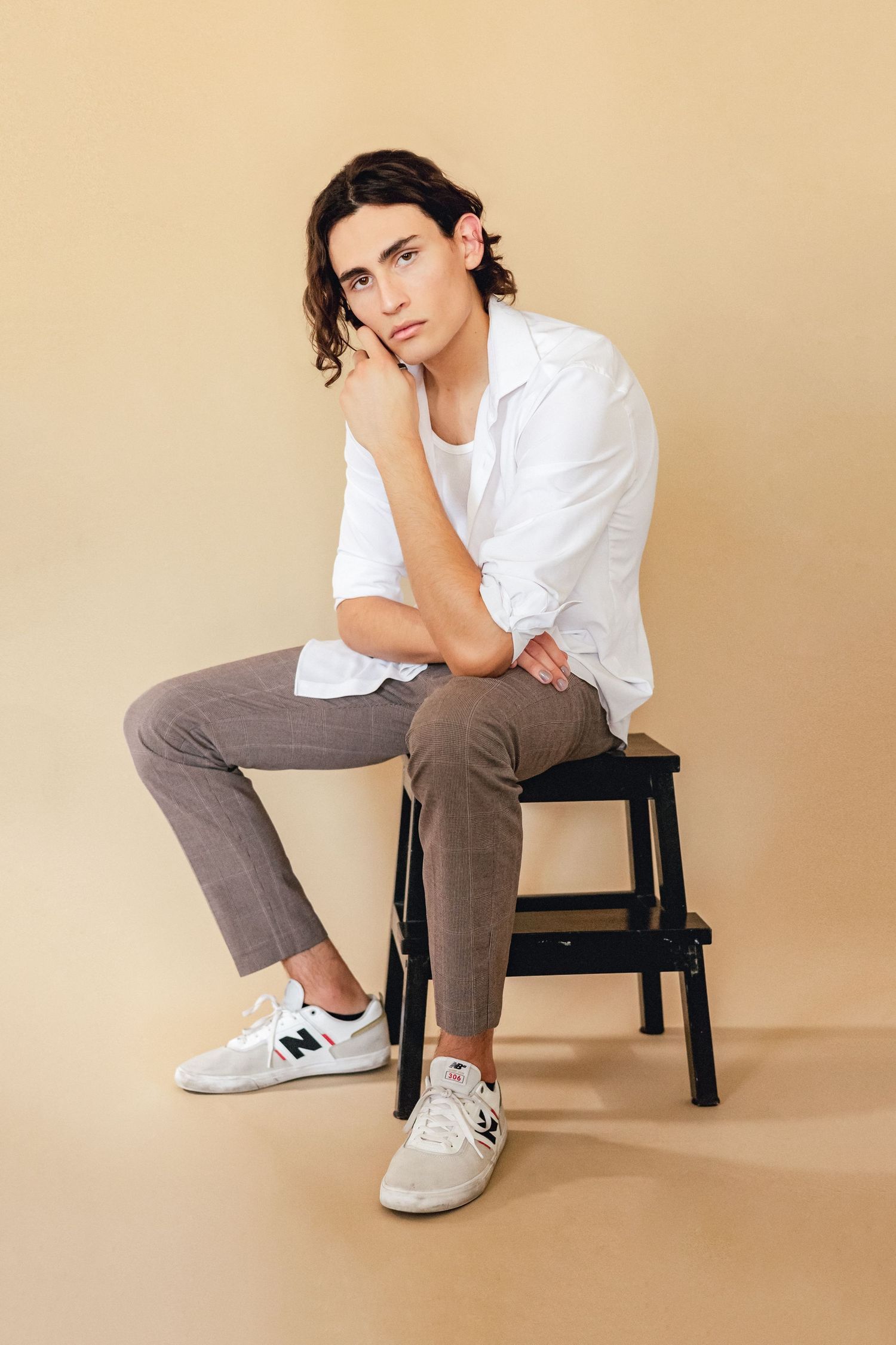 Model in white shirt and brown pants sits thoughtfully on black stool against beige backdrop wearing casual sneakers.
