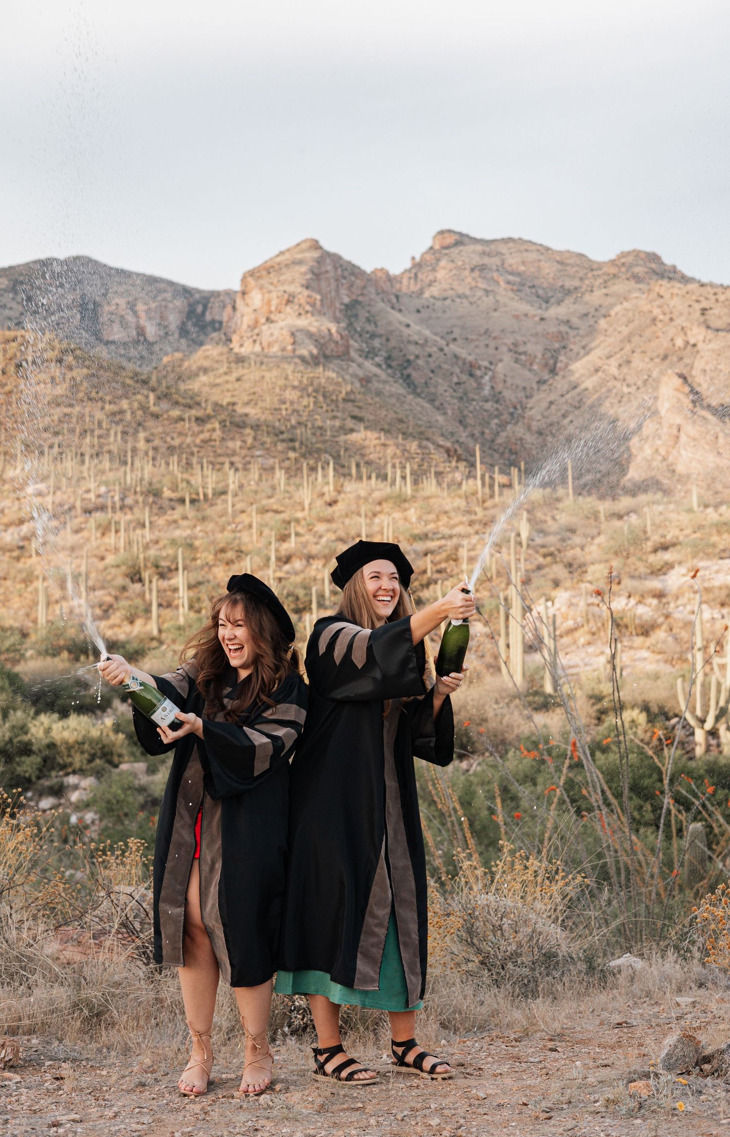 Two graduates celebrate with champagne bottles in the desert with mountains and saguaro cacti in the background.