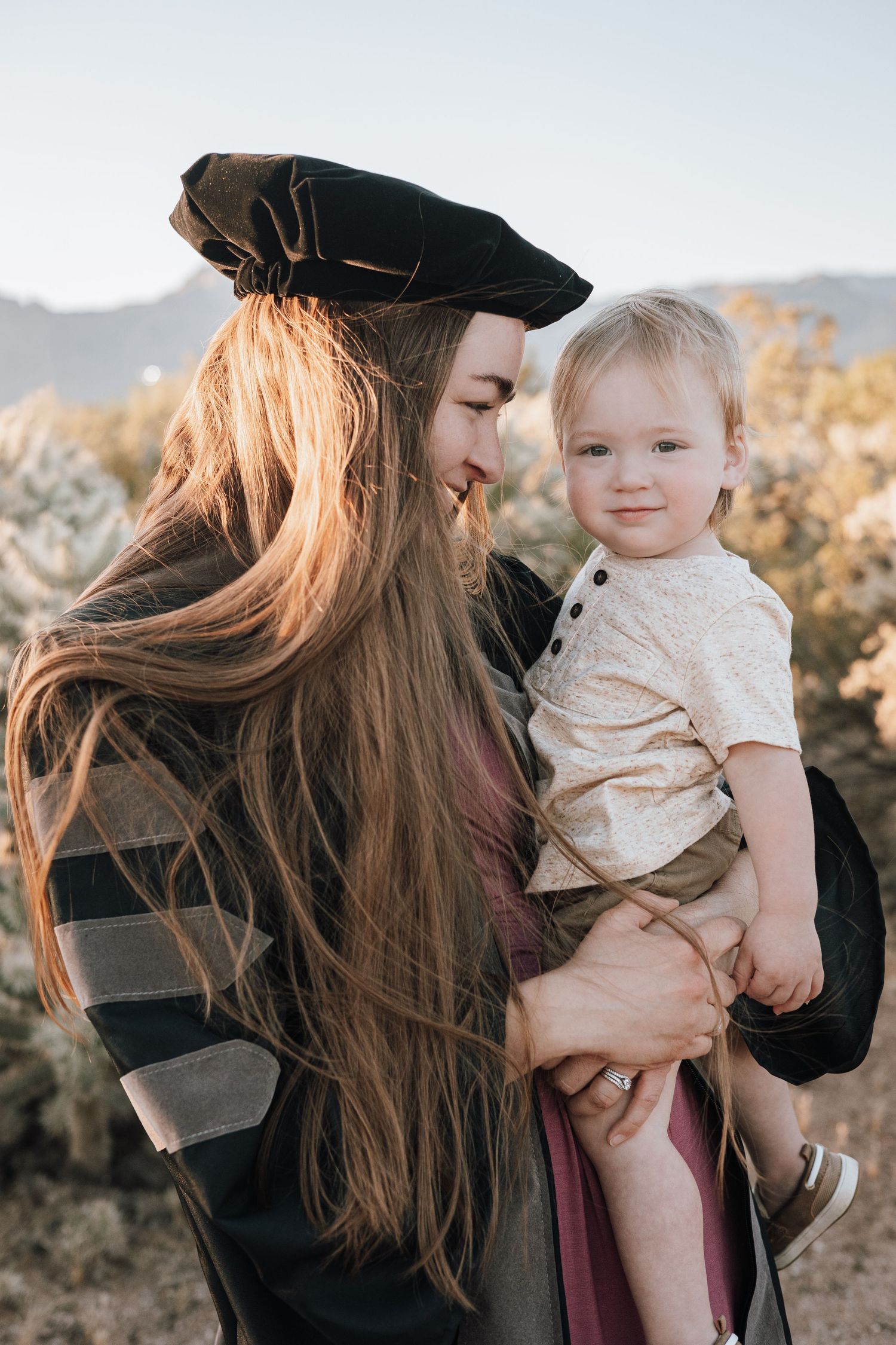 A parent with long flowing hair wearing a black hat holds a small child in casual attire against a sunset mountain backdrop.