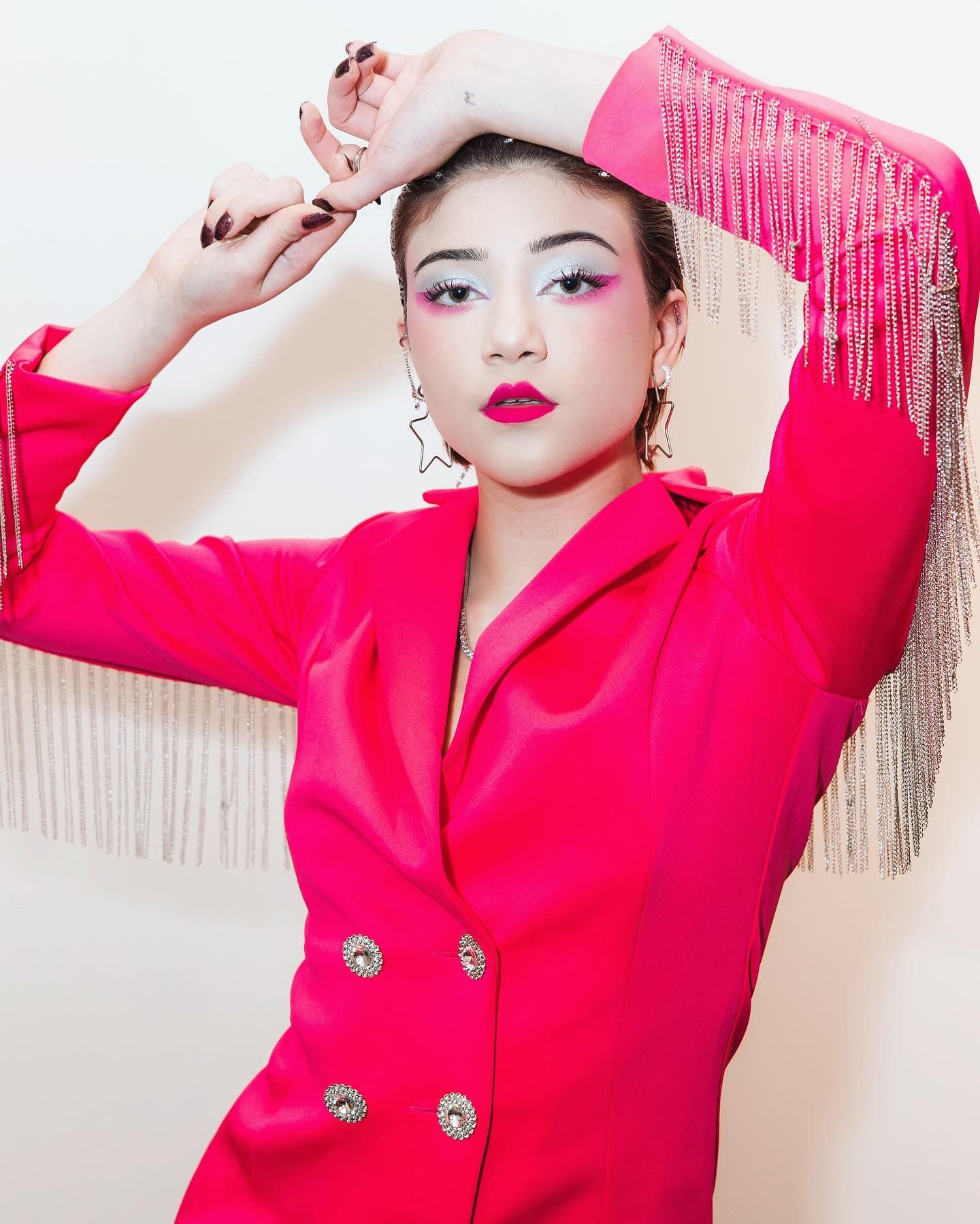 A model in vibrant pink blazer dress with statement makeup and silver fringe backdrop poses with raised arms.
