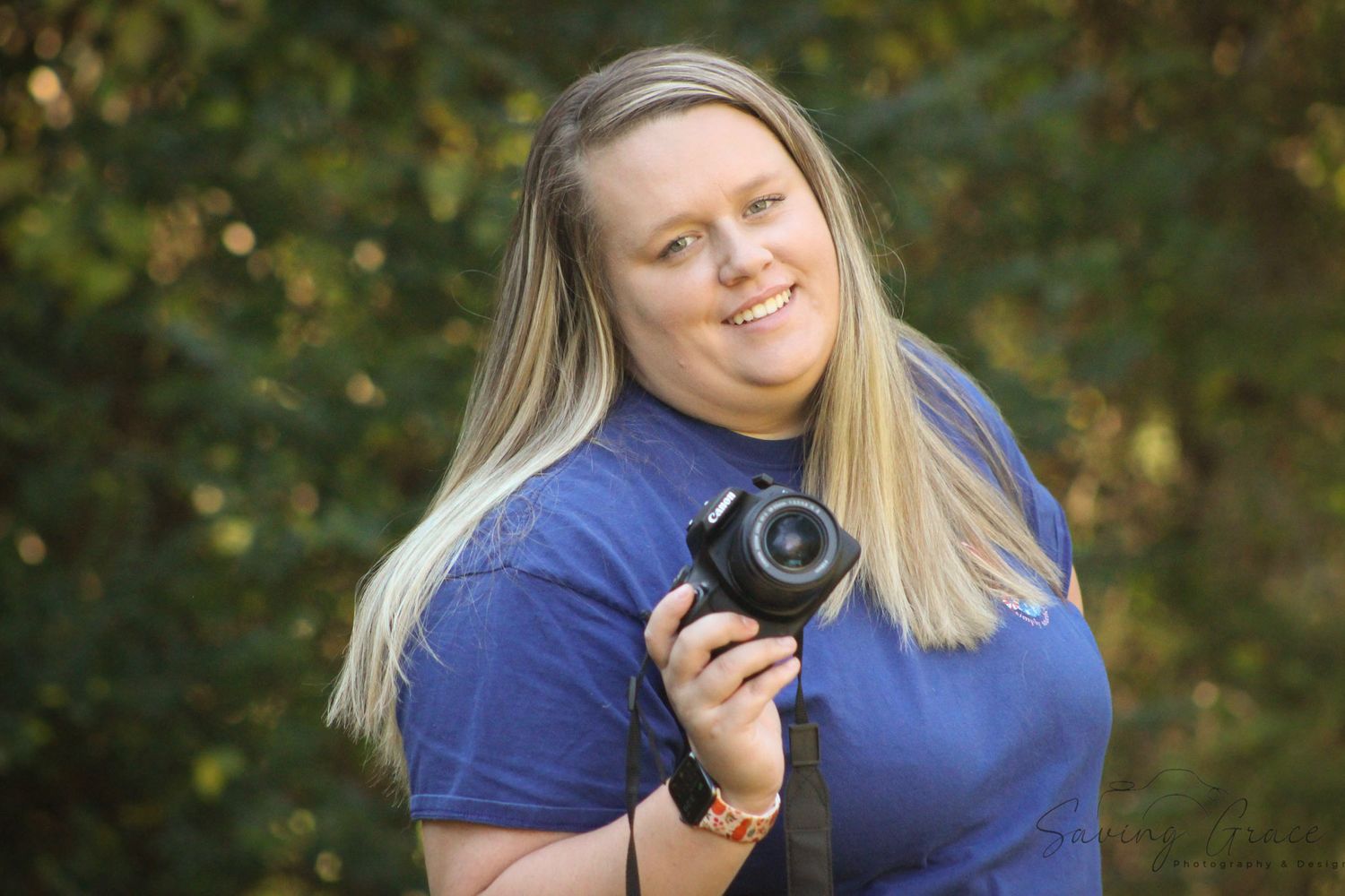Professional photographer in blue shirt smiling while holding camera outdoors with natural background.
