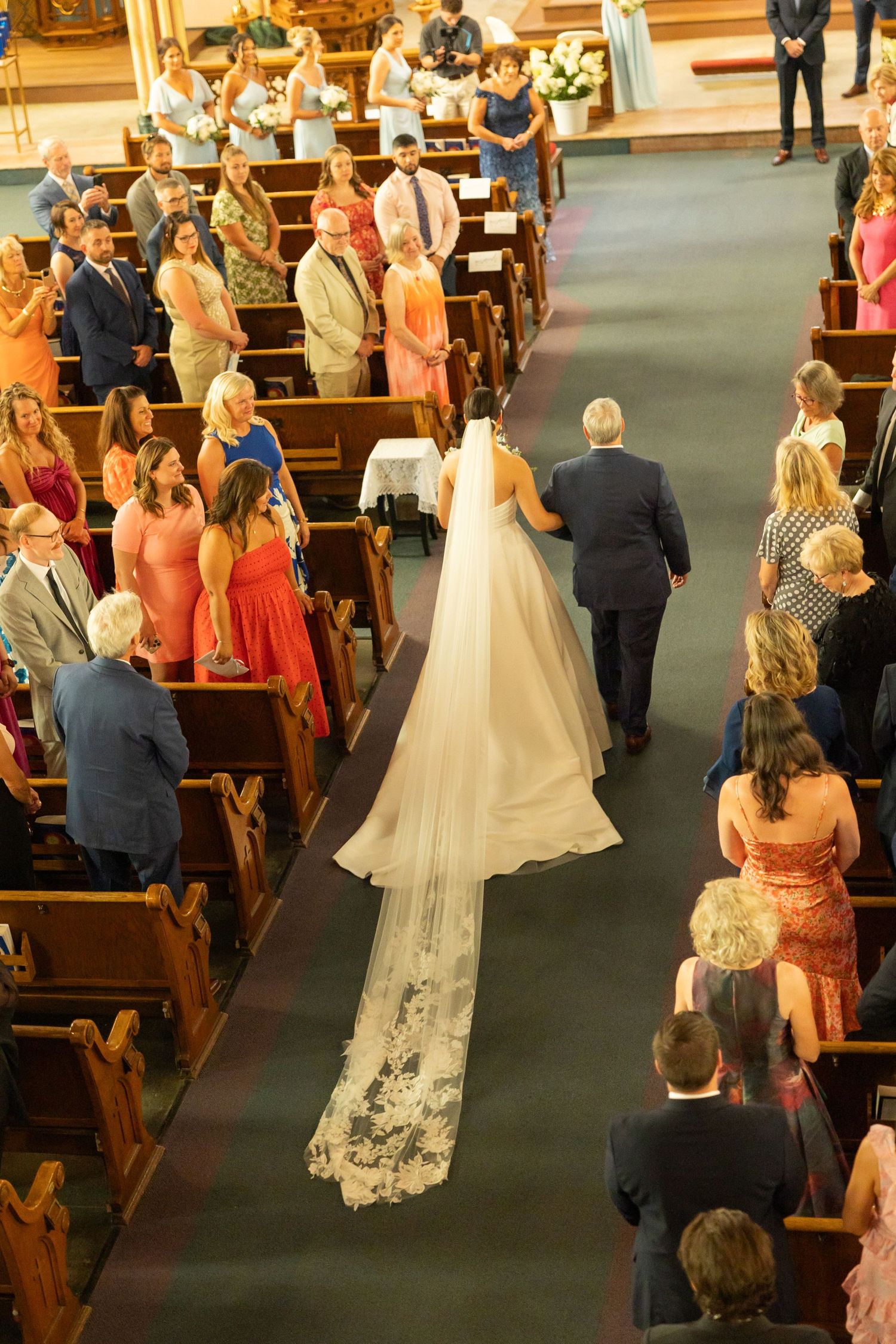 Bride in flowing lace veil walks down church aisle during wedding ceremony as guests watch from wooden pews.