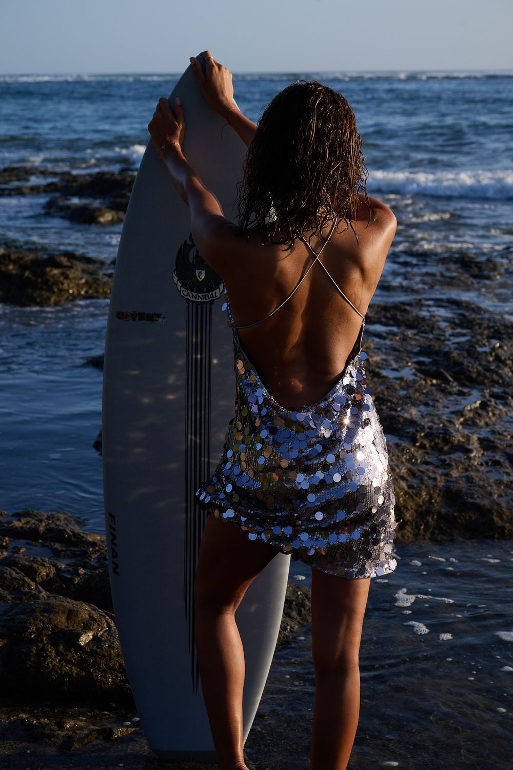 A surfer in floral swim shorts stands along rocky coastline with surfboard at sunset near ocean waves.