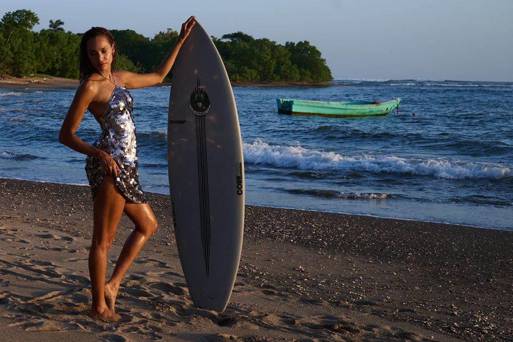 A surfer with a surfboard stands on a dark sand beach at dusk with a small boat visible in the ocean background.