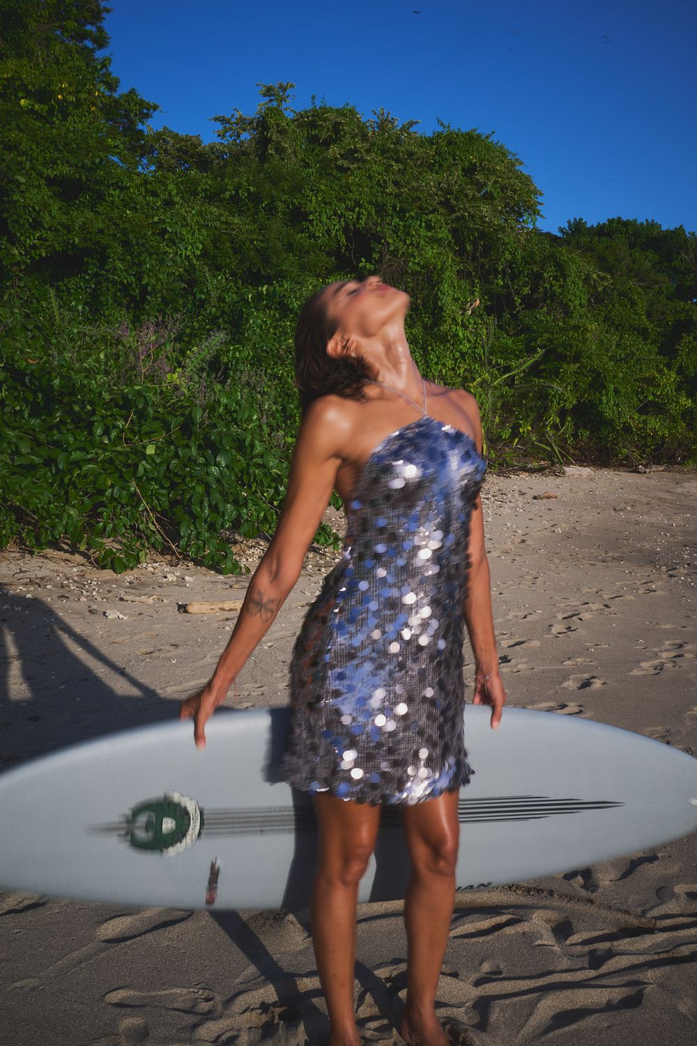 A silver sequin dress sparkles on a sunny beach with a surfboard visible in the background.