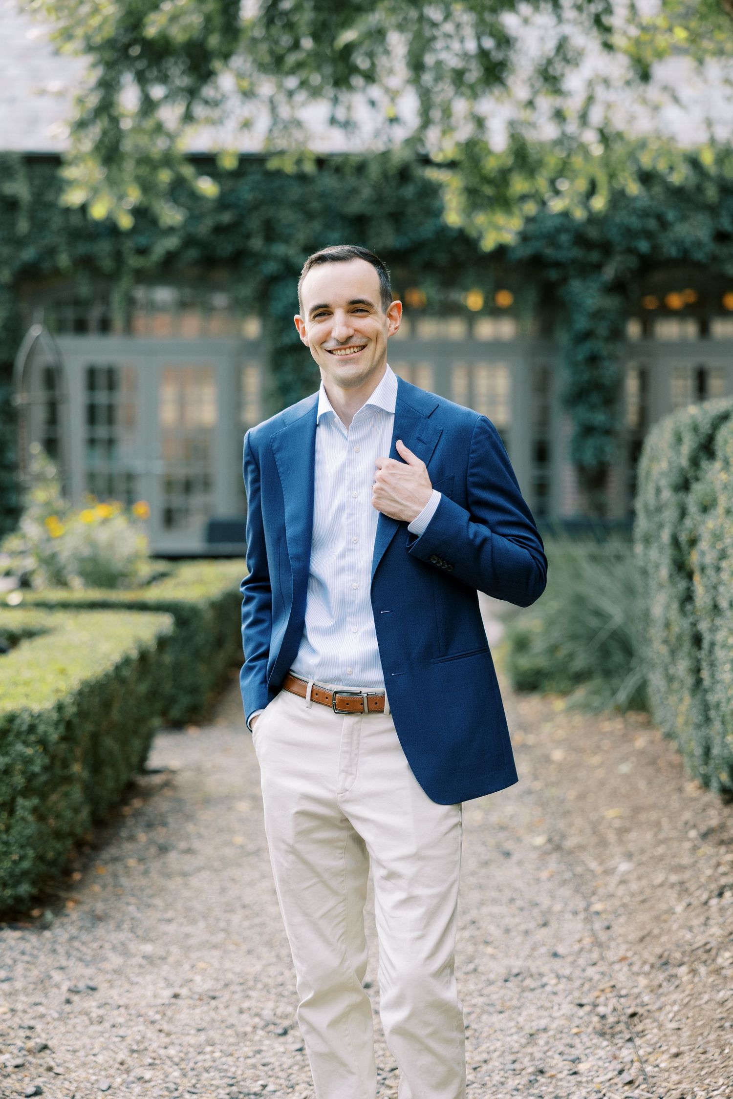 A groom in a navy blazer, white pants and shirt poses confidently on a garden path during a wedding photoshoot at Ninety Acres.