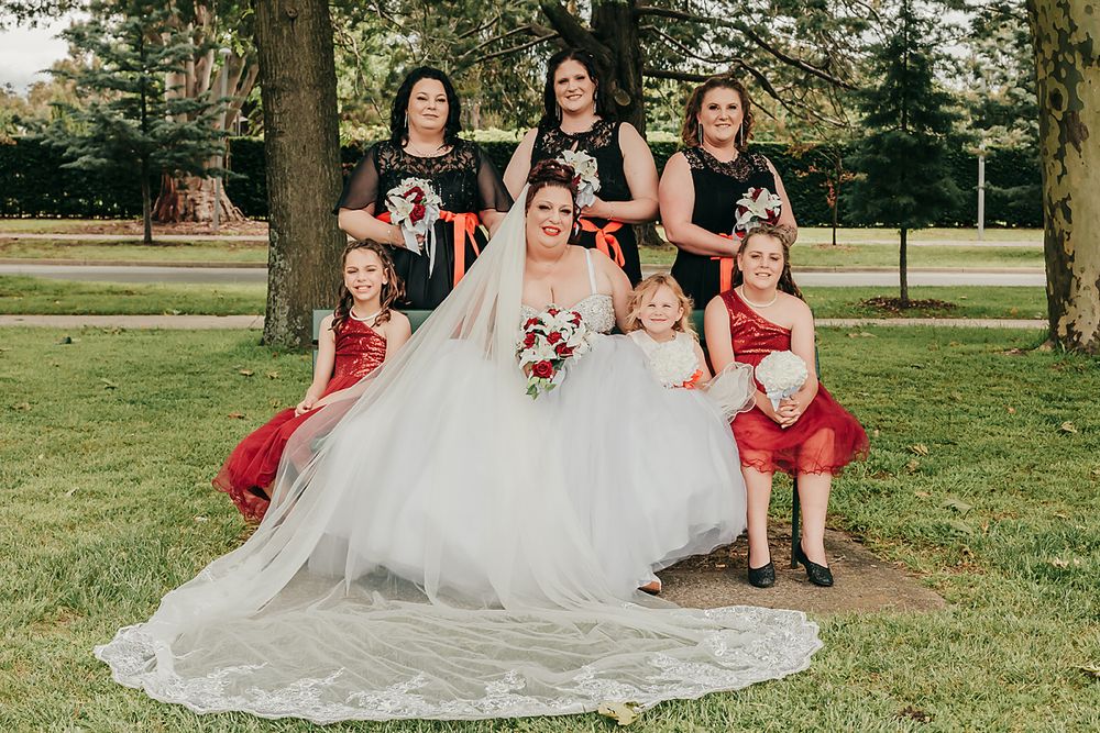 Wedding party members in red dresses and suits pose together on grass during outdoor summer celebration.