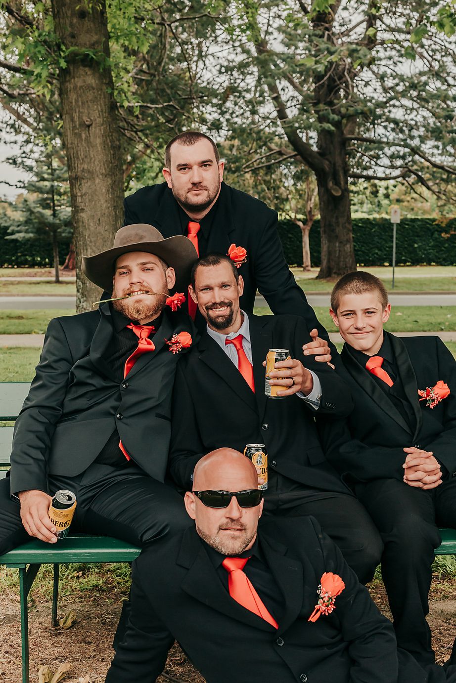 Groomsmen in black suits with orange ties and boutonnieres pose casually on bench outdoors with drinks in hand.